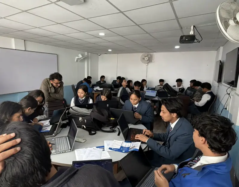 A packed computer lab with students in uniform working at laptops while an instructor helps at one of the tables.