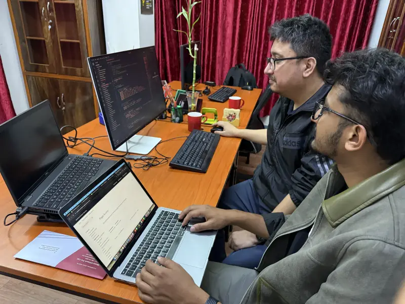 Two developers working side by side at a desk, a code editor open on the monitor and a laptop in the foreground.