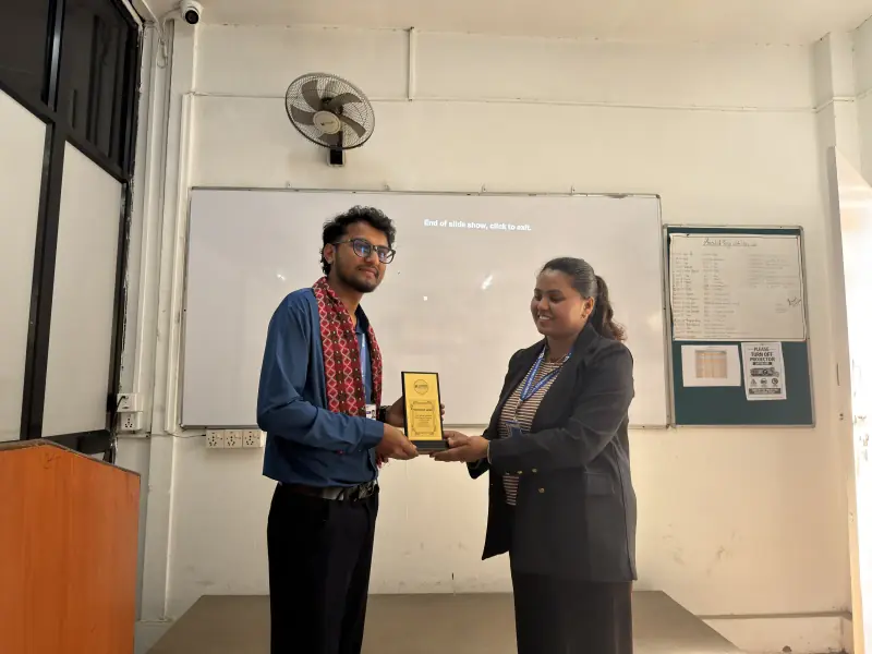 Two people holding a small engraved plaque between them at the front of a classroom.